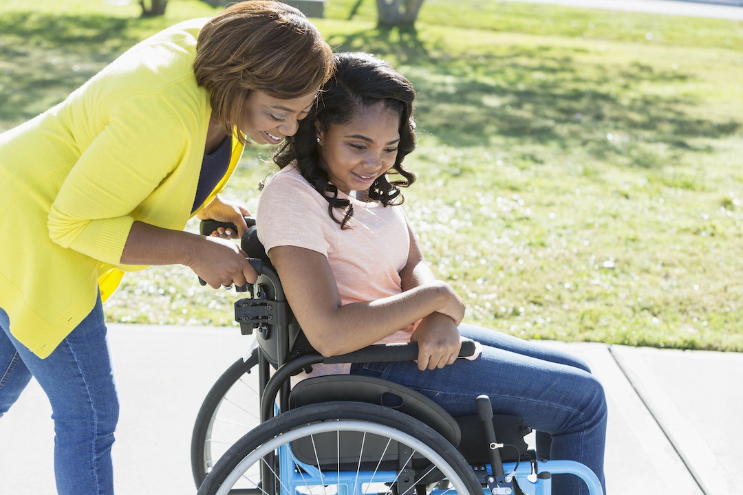 A carer assisting a young woman in a wheelchair outdoors in a park